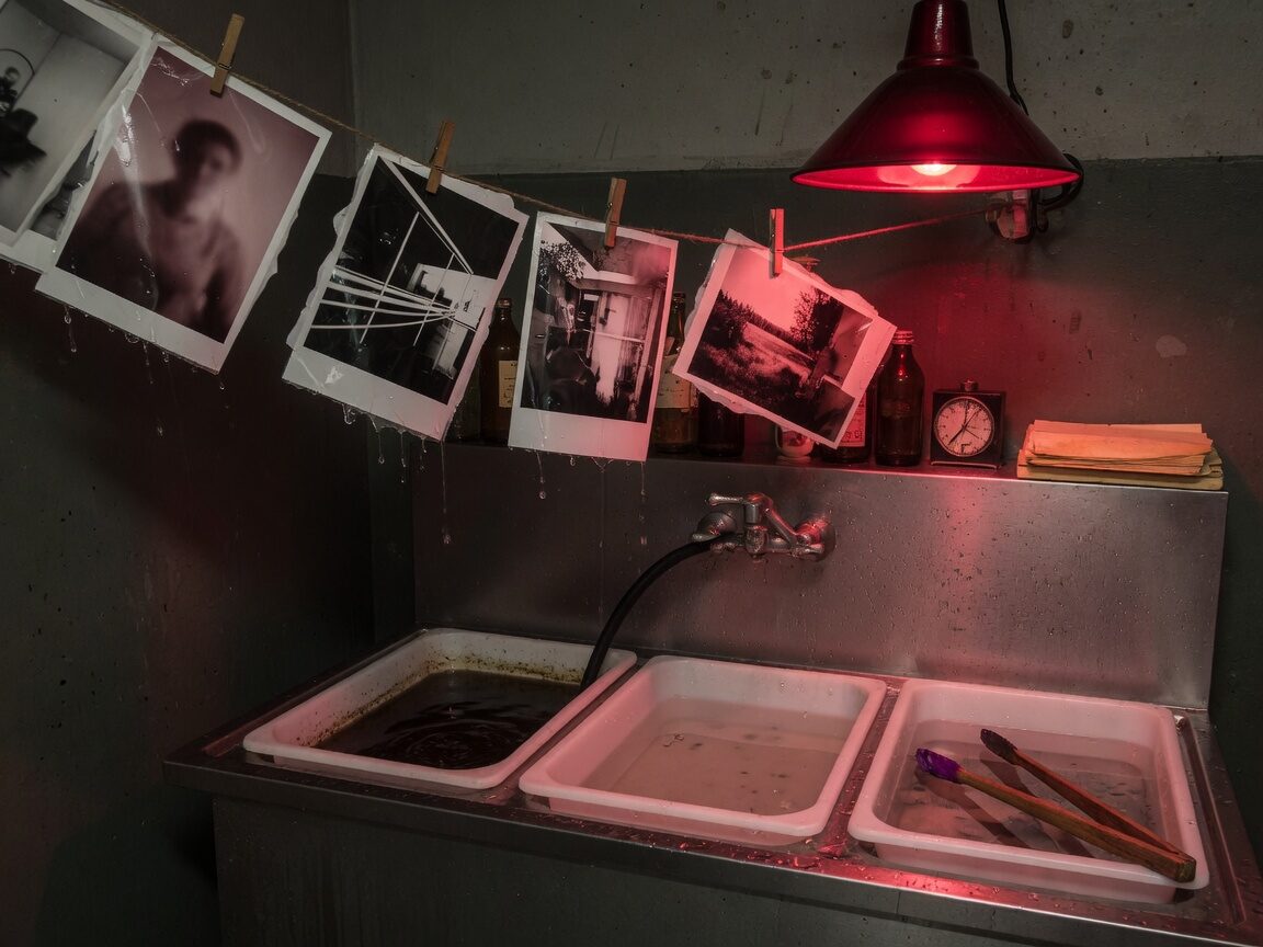 Photographic darkroom under a red safelight, developing trays and wet prints on a line.