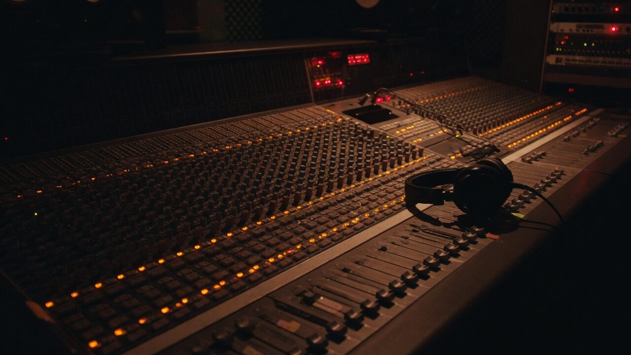 Mixing desk and headphones in a dim control room, small amber and red LEDs.